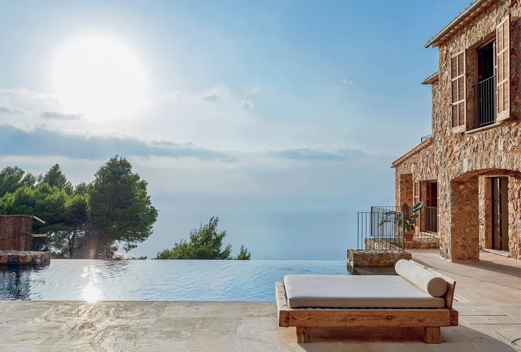 This image presents two distinct views of a luxurious villa, each showcasing its unique features. In the top half, an infinity pool dominates the scene, stretching from left to right and beyond the frame's edge. The pool is surrounded by lush trees, adding a touch of natural beauty to the setting. Meanwhile, in the bottom half, the villa itself takes center stage, featuring stone walls with numerous windows and a door. A pair of sun loungers are positioned on the patio outside the villa, inviting relaxation and leisure. The image's background sets the tone for an idyllic getaway, with its serene atmosphere and picturesque surroundings evoking feelings of tranquility and luxury.