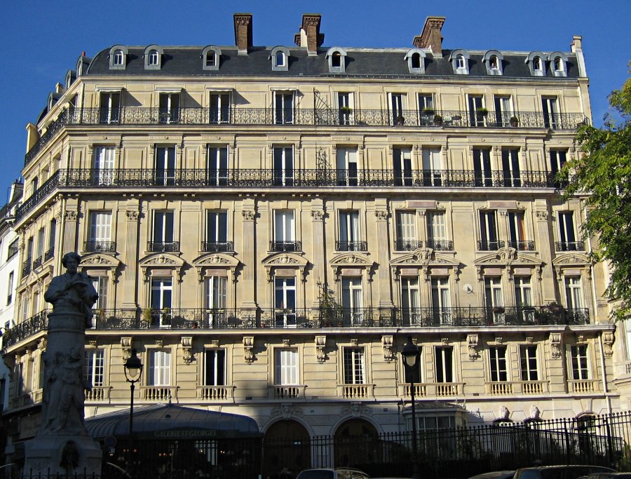 The image depicts a large, multi-story building with a cream-colored facade and numerous windows. The building features ornate details such as balconies, railings, and intricate stonework. * **Building:** * Cream-colored facade * Multiple stories * Numerous windows * Ornate details: * Balconies * Railings * Intricate stonework * **Background:** * Clear blue sky The image showcases a grand, historic building with elegant architectural features set against a serene backdrop.