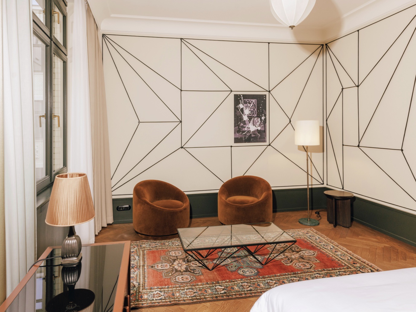This photograph captures a serene bedroom, with the bed situated in the bottom-right corner and its edge cropped out of view. The room's walls are adorned with an intricate black geometric pattern on white wallpaper, complemented by two brown chairs positioned against one wall. A small table stands to the left of the chairs, while a floor lamp is placed to their right. The wooden floor is partially covered by an ornate rug featuring a red, tan, and green floral design. In the foreground, a black glass-topped coffee table sits atop another patterned rug, which adds depth and texture to the space. The overall ambiance of the room exudes warmth and coziness, creating a peaceful retreat for relaxation and rest.