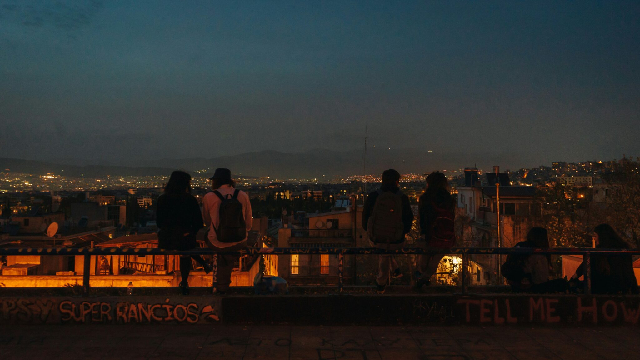 The image depicts a group of people standing on a rooftop, overlooking the city below, with graffiti and writing covering the walls. The scene is set against a backdrop of a darkening sky. * A group of people are standing on a rooftop: + They are facing away from the camera. + Their silhouettes are visible against the fading light. * The city lights can be seen in the background: + The lights twinkle like stars. + The cityscape stretches out as far as the eye can see. * There is graffiti on the walls: + The writing is illegible due to its faded state. + It appears to be a mix of colors and fonts. The image suggests that the group has gathered on the rooftop to take in the view, perhaps at sunset or dusk. The presence of graffiti adds a layer of urban grittiness to the scene, highlighting the contrast between the natural beauty of the cityscape and the raw energy of the people who call it home.