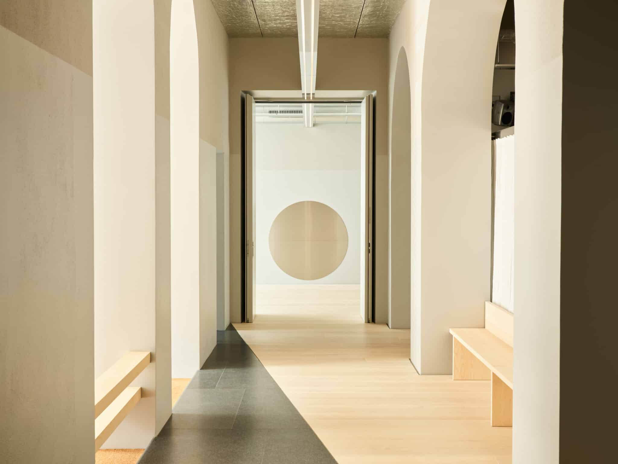 This photograph captures a serene and minimalist hallway, characterized by its white walls, arched doorways, and pale wood flooring. The hallway is set against a backdrop of stark, featureless white walls, punctuated by three arches that lead to various areas within the space. The central doorway frames a striking tan circle on the wall behind it, while another doorway to the right appears to be closed off with what resembles a piece of wood or drywall. The pale wood flooring adds warmth and texture to the otherwise monochromatic setting, creating a sense of balance between light and shadow. Natural light pours in from the left side of the image, illuminating the space and casting subtle shadows on the walls. Overall, this photograph presents a peaceful and contemplative atmosphere, inviting viewers to immerse themselves in its simplicity and elegance.