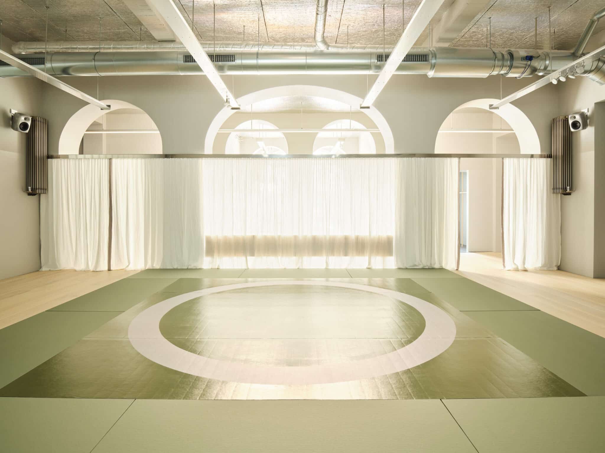 This image depicts a spacious, well-lit room with a large circular mat in the center, surrounded by three arched openings on the back wall. The light-colored wood floor and green mats create a neutral atmosphere, while white curtains adorn the arches. The ceiling features exposed pipes and ductwork, suggesting an industrial setting. Two speakers mounted on either side of the room add to the ambiance. Overall, the image exudes a sense of calmness and serenity, evoking the feeling of being in a peaceful environment.