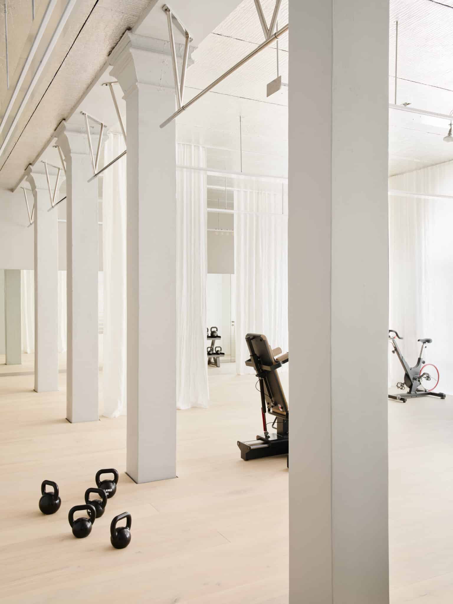 Alt tag: "Bright and spacious modern gym interior featuring white columns, kettlebells on the floor, and exercise equipment including a stationary bike and an adjustable bench."