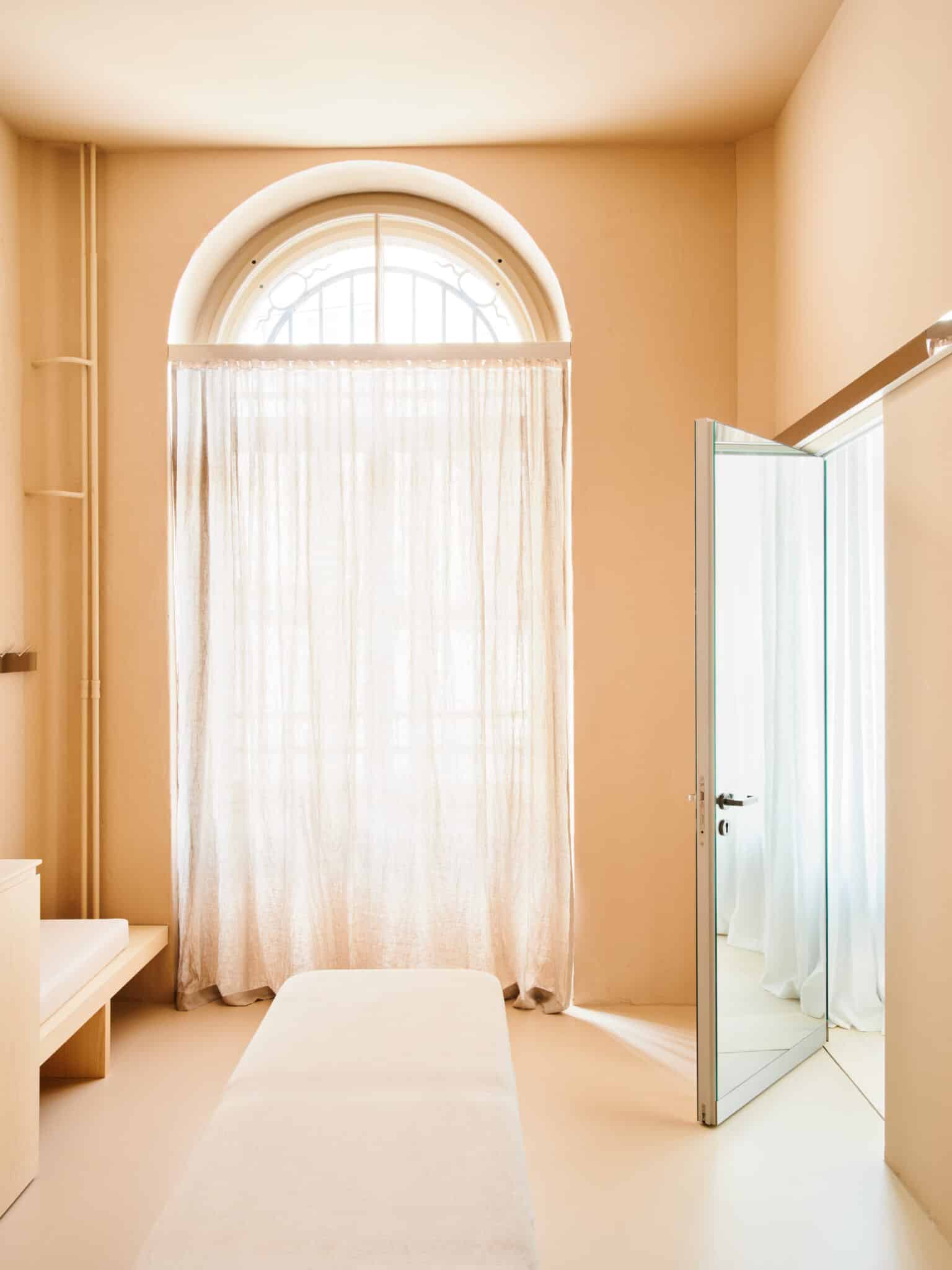 Alt tag: "Minimalist interior of a sunny room featuring a large arched window with sheer curtains, a light-colored bench, and a partially open door reflecting soft light."