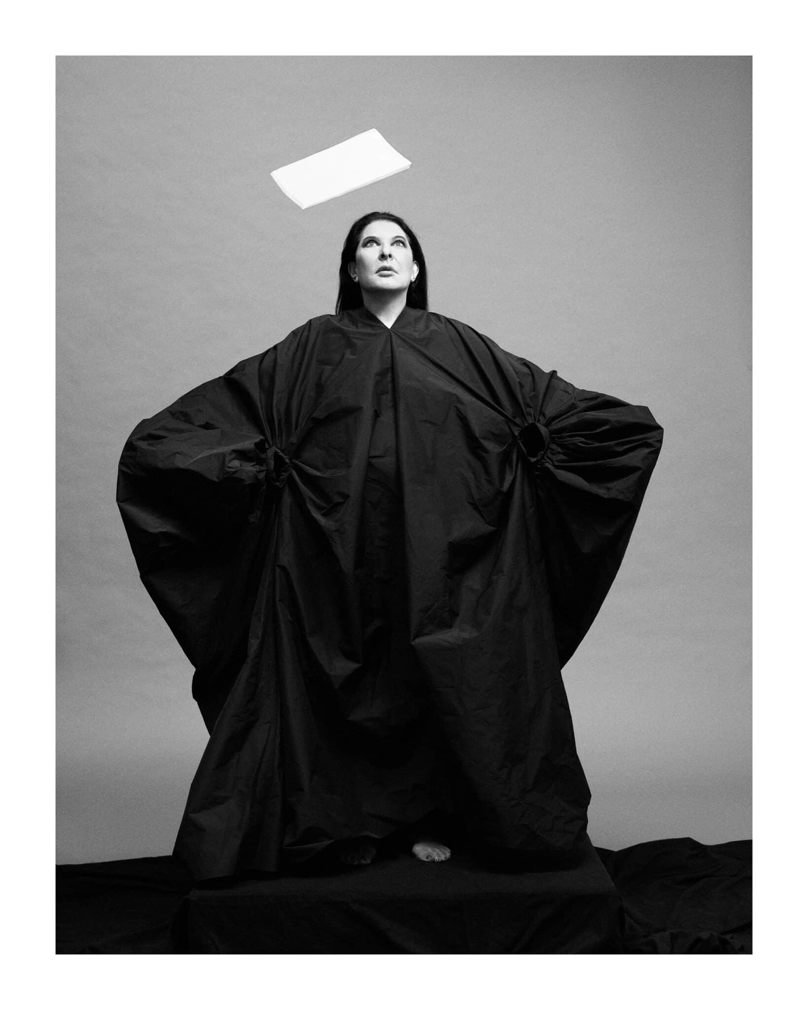 The image is a black and white photograph of a woman in a long robe standing on a small platform, with her hands in the pockets of the robe. The background is out of focus. * **Woman:** * Standing on a small platform * Wearing a long robe that covers her body * Her hands are in the pockets of the robe * Looking up at something above her head * **Background:** * Out of focus * Appears to be a room or building The overall atmosphere of the image is one of contemplation and introspection, as if the woman is lost in thought. The use of black and white photography adds to the somber mood, emphasizing the simplicity and elegance of the subject's attire.