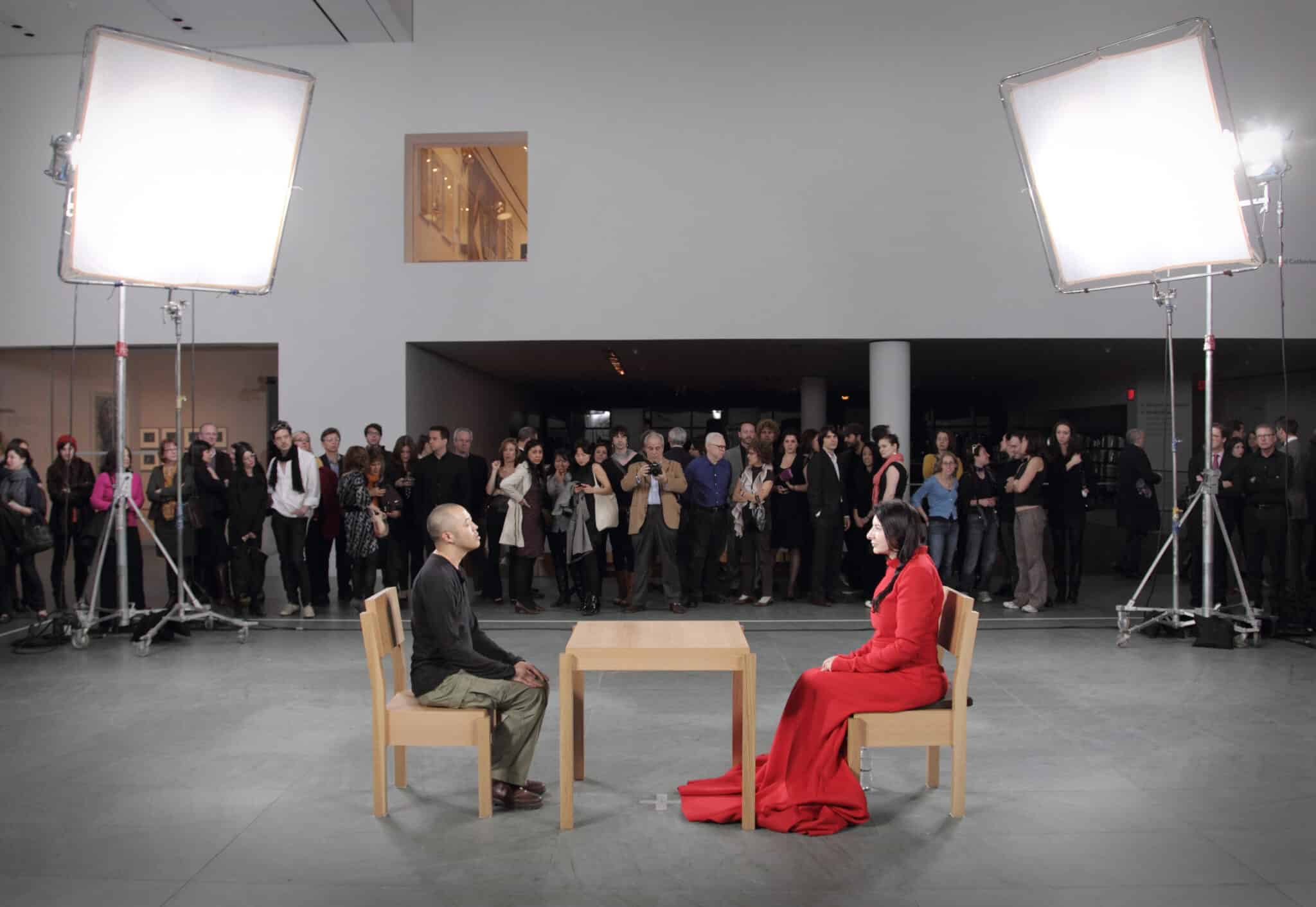 This image captures a moment from the filming of an interview, with two individuals seated in chairs facing each other. The man, dressed in a black shirt and khaki pants, sits on the left side of the frame, while the woman, wearing a vibrant red dress, occupies the right side. A small wooden table is positioned between them. The room is well-lit, with natural light pouring in through windows and additional lighting from large white panels suspended above the subjects. The surrounding space is filled with an audience, comprising both men and women, who are observing the interview taking place.