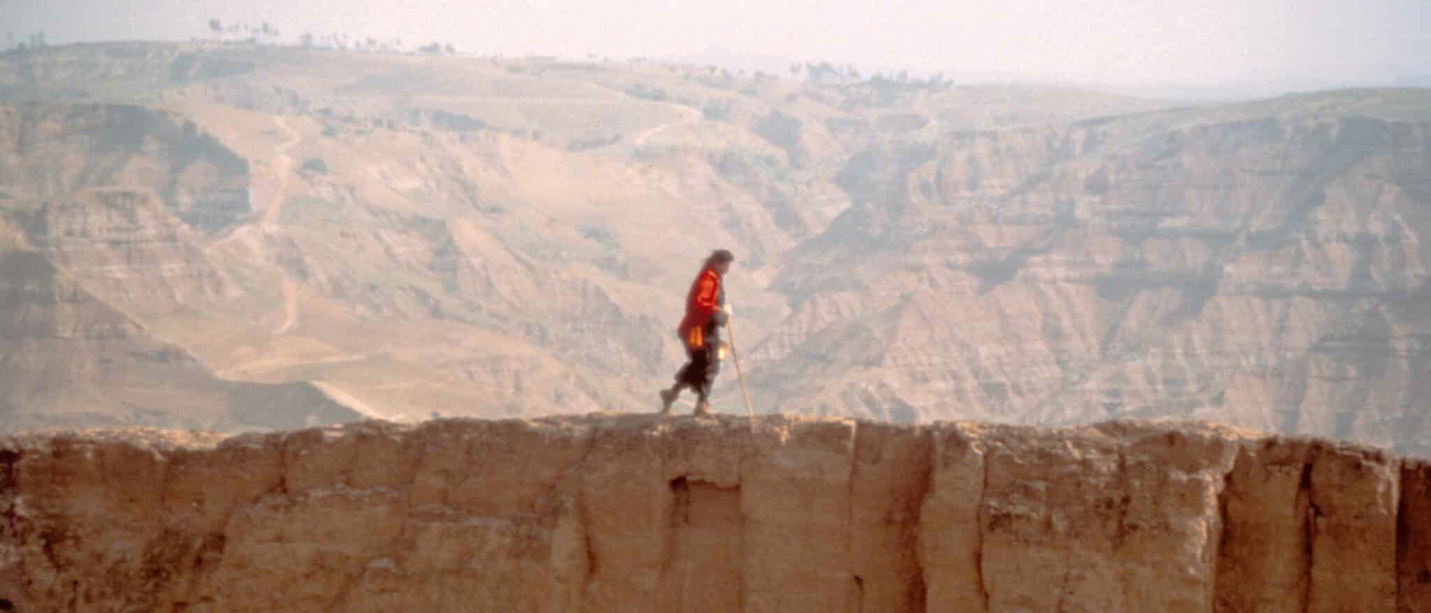 A lone hiker in a red jacket walking along a rugged cliff edge, with vast, arid mountains and valleys in the background.