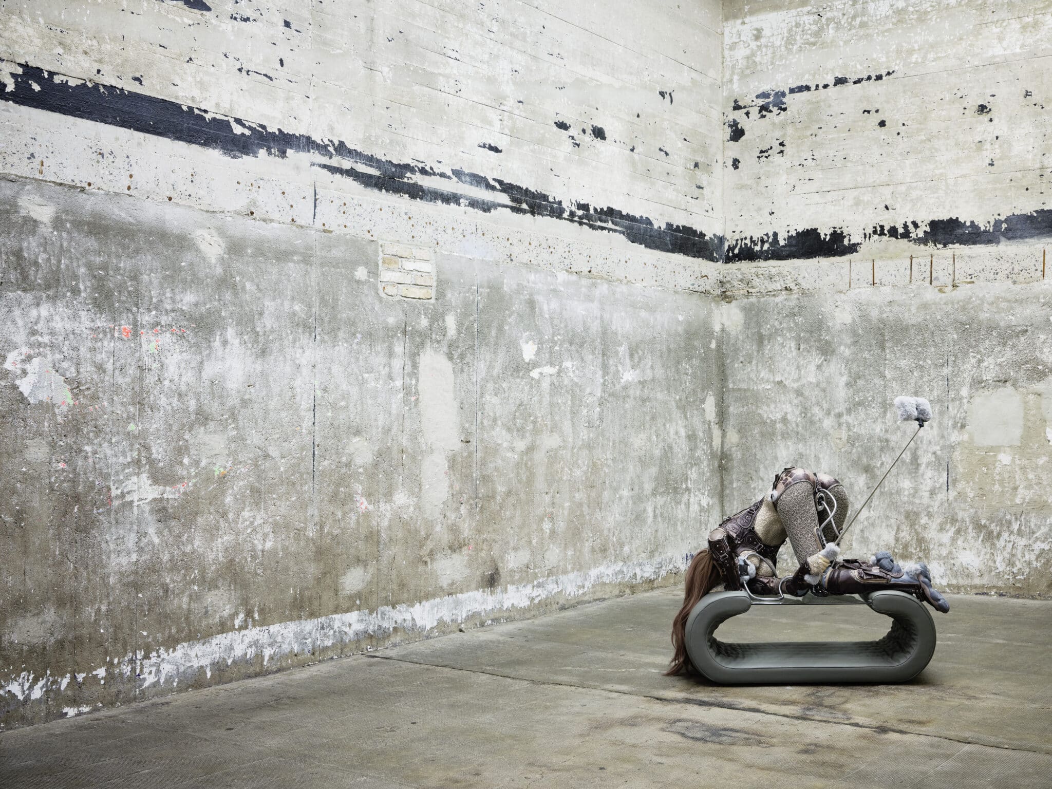 A figure in elaborate costume rests on a modern, curved bench in an industrial space with textured concrete walls, blending themes of art and surrealism.