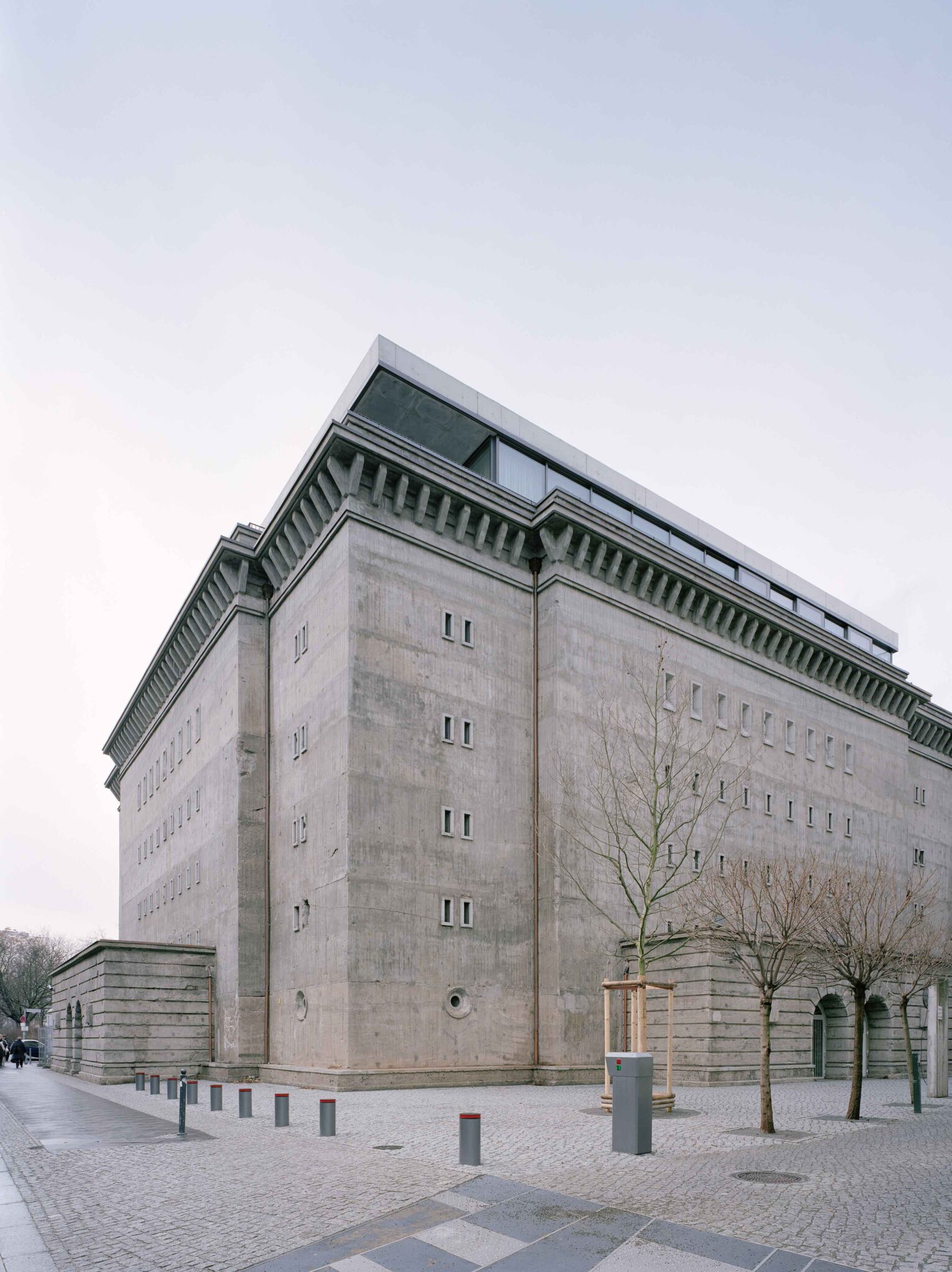 Alt tag: "View of a modern concrete building with a flat roof and geometric design, surrounded by a pedestrian area featuring trees and street elements."