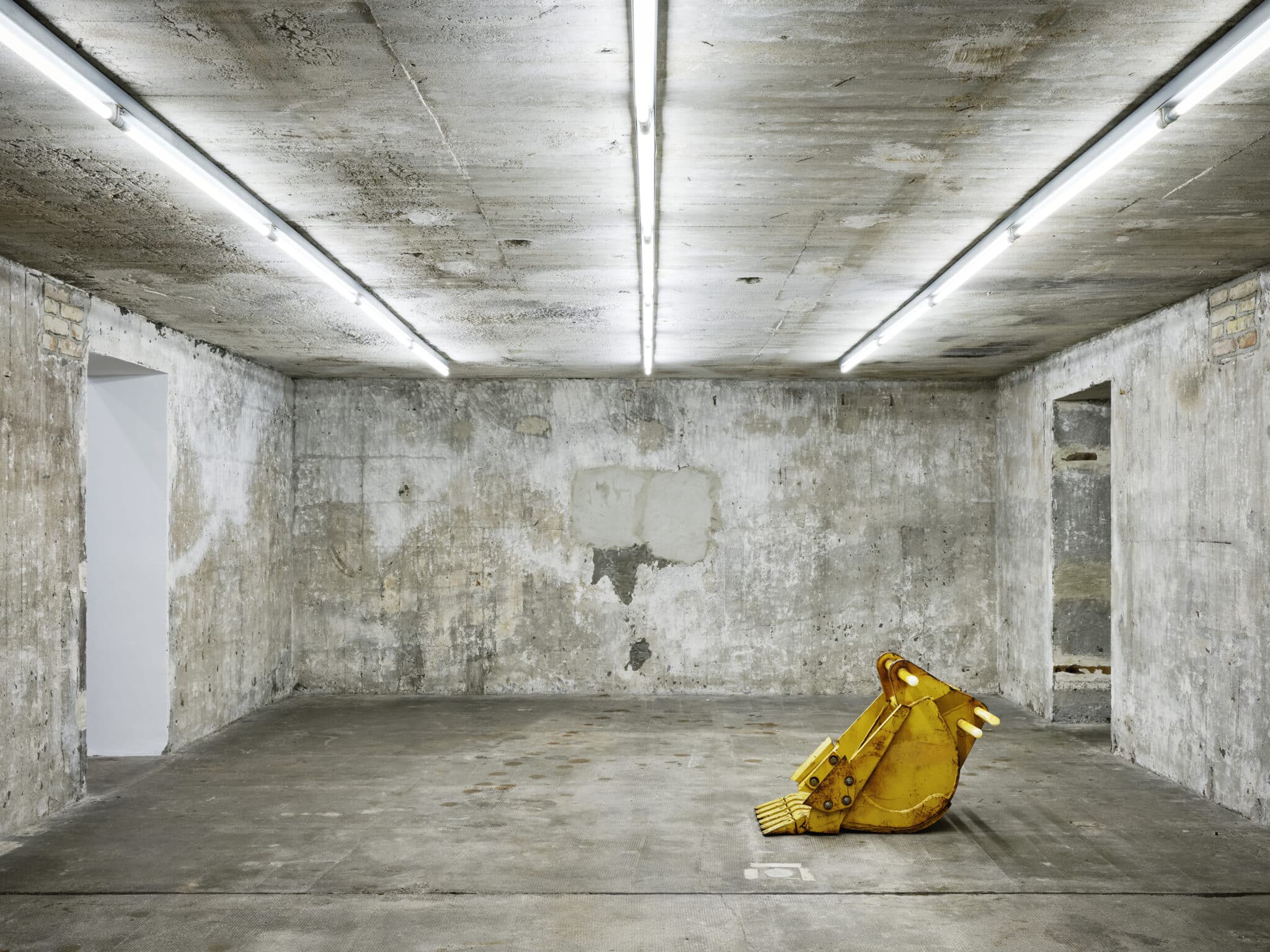 This photograph features a striking yellow metal excavator bucket situated in the center of a concrete room, surrounded by white fluorescent lights that illuminate the ceiling and walls. The wall to the left is partially visible, with a small opening leading to another space. The bucket's bright color provides a stark contrast against the otherwise drab and weathered background, creating an intriguing visual effect.