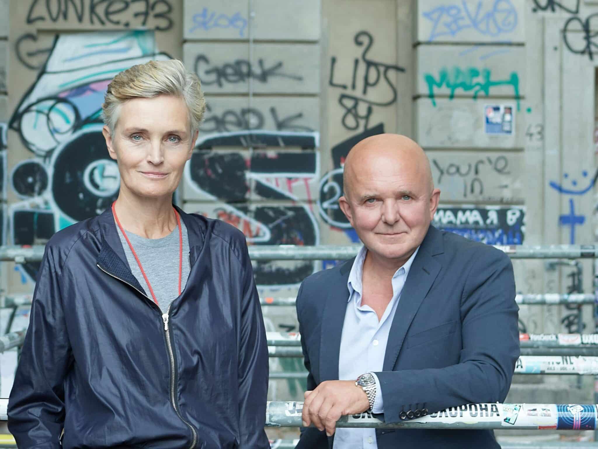 A man and a woman pose confidently in front of a graffiti-covered wall; the woman wears a navy jacket and has short blonde hair, while the man is bald and dressed in a dark blazer over a light shirt.