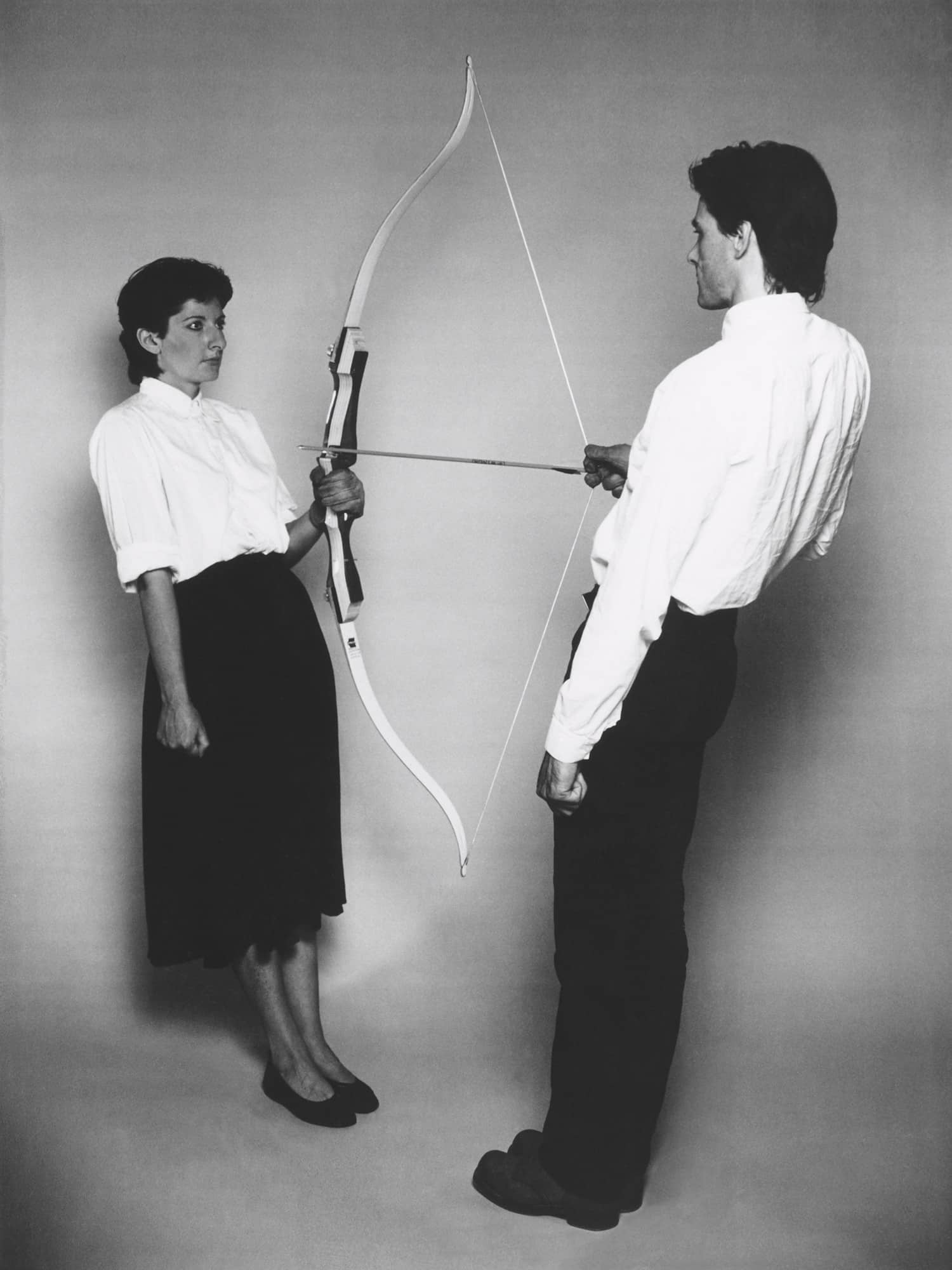 A man in a white shirt aims an unstrung bow at a woman in a white blouse and black skirt, who holds the bowstring with a concerned expression, in a dramatic black and white composition.