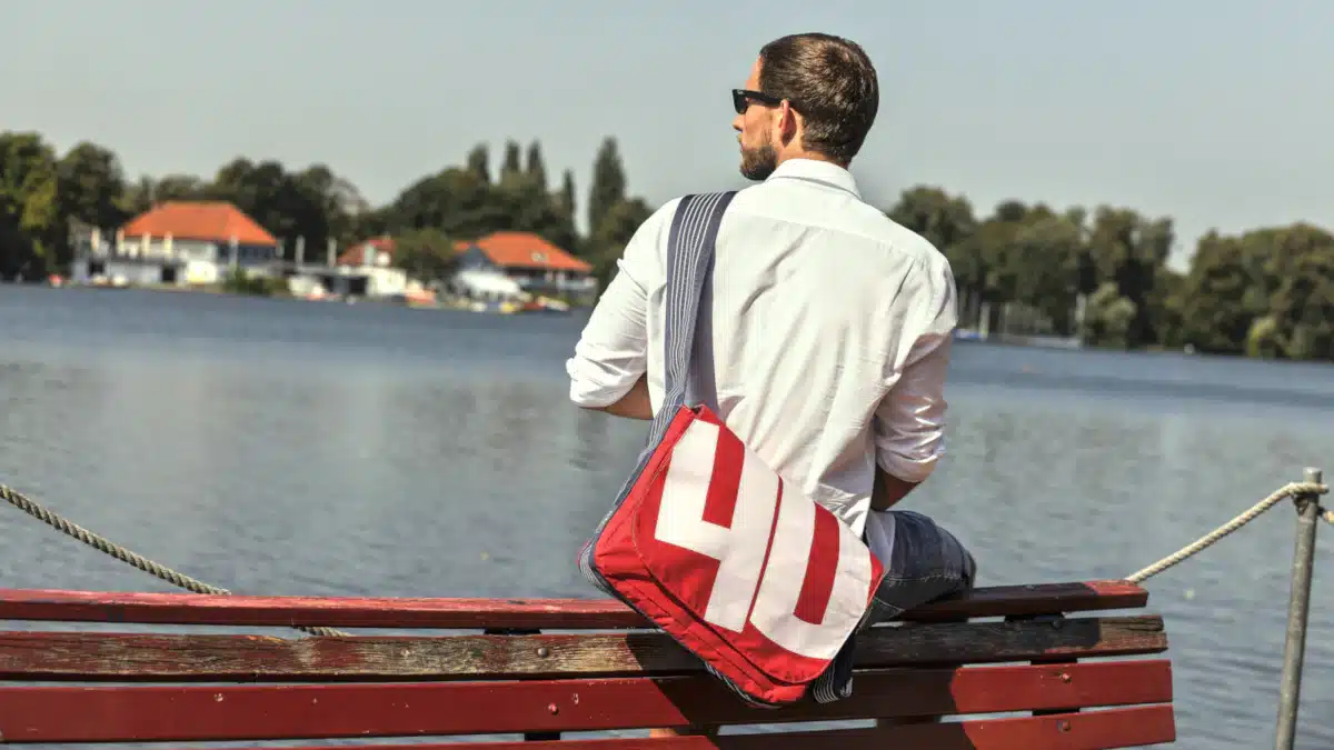 The image depicts a man sitting on the edge of a wooden boat or dock, gazing out at the water. He is dressed in a white shirt and has short brown hair, with his back turned towards the camera. * **Man's Appearance** * The man wears a white dress shirt. * His hair is short and brown. * **Bag** * A red bag hangs over his shoulder. * It features large white letters that read "YO". * **Boat or Dock** * The man sits on the edge of a wooden boat or dock. * There are ropes hanging down from the boat to the water below. * **Water and Background** * In the background, there is a body of water with trees in the distance. * A few buildings can be seen behind the trees.
