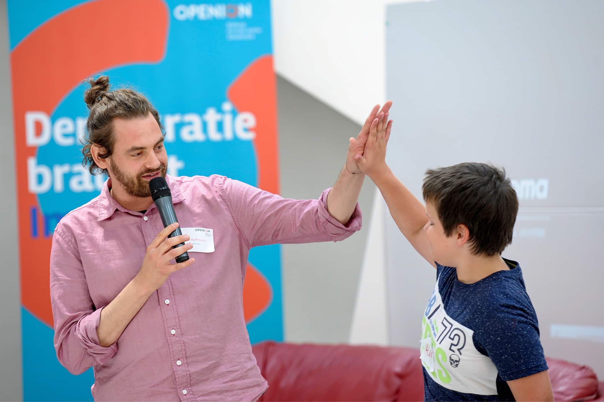A man speaking into a microphone gives a high five to a young boy, both smiling, in front of a colorful banner that promotes democratic engagement.